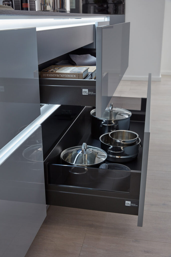 Offene Küchenschubladen mit Metalltöpfen und einem Stapel Bücher darin, vor einem eleganten, modernen Design.