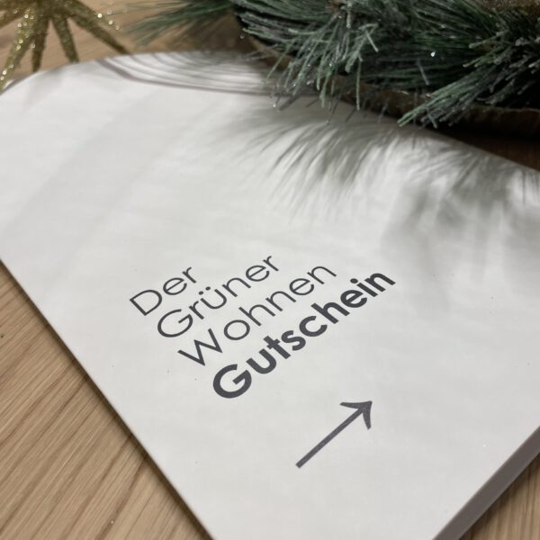 Geschenkkartenumschlag auf einer Holzoberfläche mit dem Text „Der Grüner Wohnen Gutschein“ und einem Pfeil. Oben Tannenzweige.