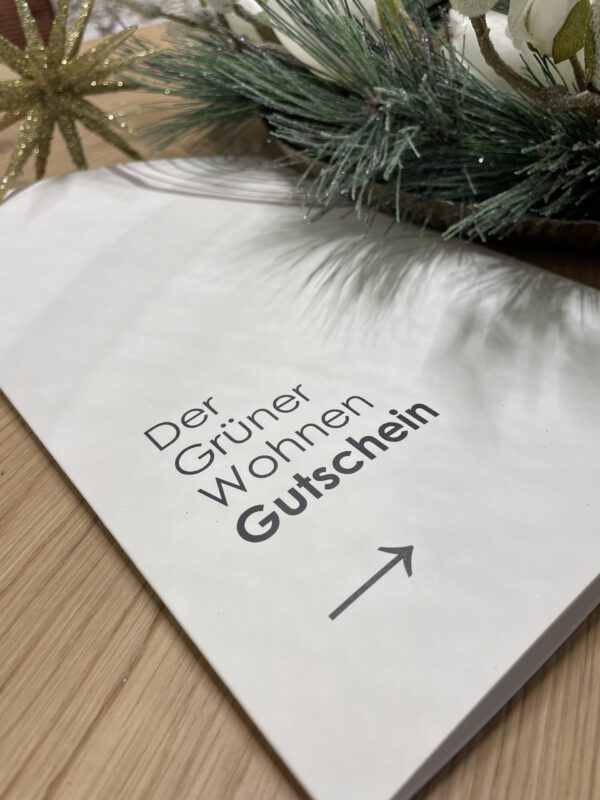 Geschenkkartenumschlag auf einer Holzoberfläche mit dem Text „Der Grüner Wohnen Gutschein“ und einem Pfeil. Oben Tannenzweige.
