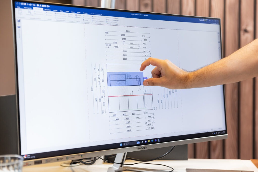 Eine Person zeigt auf eine Küchenplanung, die auf einem großen Computermonitor in einer Büroumgebung angezeigt wird.
