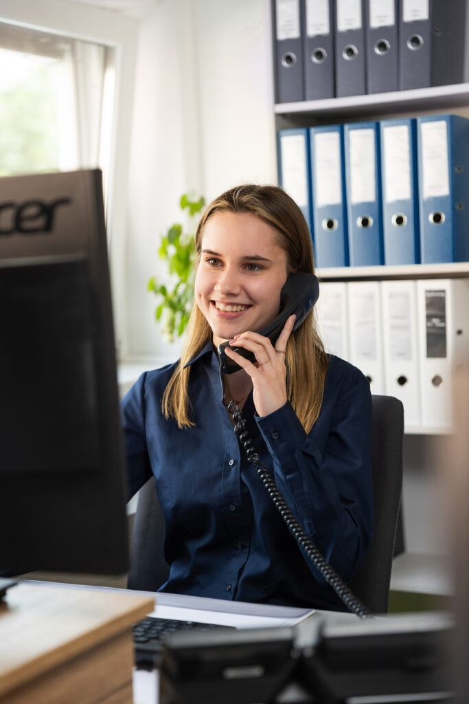 Lächelnde Frau in einem Büro, die mit einem schnurgebundenen Telefon spricht, während sie an einem Computertisch arbeitet, im Hintergrund sind Dateien.